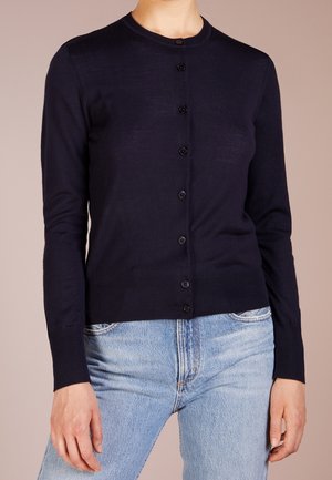 Vrouw draagt een aansluitende zwarte cardigan met knopen en lange mouwen en lichtblauwe hoge taille jeans tegen een effen achtergrond.