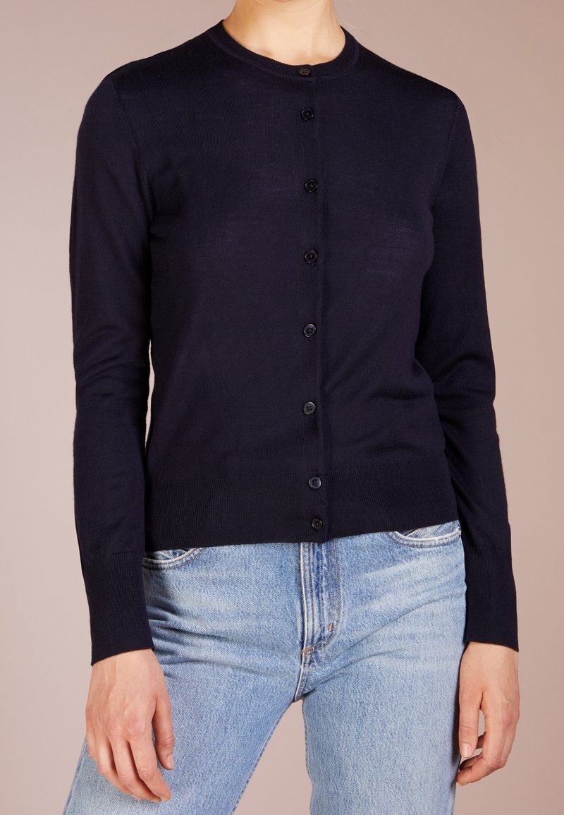 Femme portant un cardigan noir ajusté boutonné avec des manches longues et un jean bleu clair taille haute sur un fond uni.