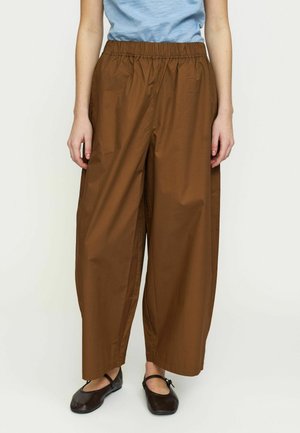 Persoană îmbrăcată în pantaloni maro largi cu talie elastică, top albastru deschis și pantofi Mary Jane maro închis, stând într-un decor simplu, cu fundal luminos.