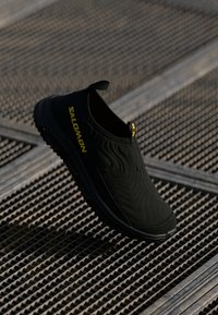 Black slip-on shoe with textured upper, featuring a wave pattern, reinforced toe, and yellow branding. Rubber sole for traction.