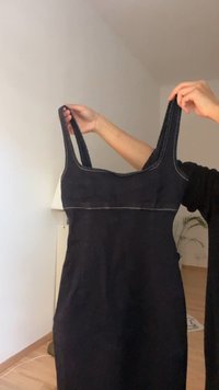Crna haljina bez rukava s ravnim dekolteom, debelim trakama i kontrastnim šavovima. Glatka tekstura, haljina srednje dužine. Minimalistički stil.