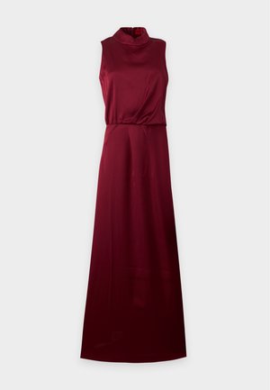 Une femme porte une robe bordeaux sans manches, longue jusqu’au sol, avec un détail drapé à nouer et des escarpins noirs, sur un fond uni.