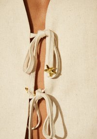 Linneöverdel i off-white med en djup V-ringning, utrustad med två knutstängningar prydda med små guldstjärnor.