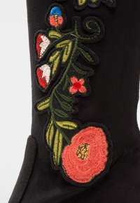 Botte en daim noir ornée d'une broderie florale vibrante en rouge, blanc et vert, avec une couture détaillée et une finition texturée.