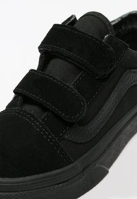Black sneakers with a textured suede upper, featuring two velcro straps and black rubber soles. Simple design with contrasting fabric details.