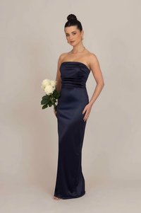 Γυναίκα που φοράει στράπλες φόρεμα από ναυτικό σατέν, κρατώντας ένα μπουκέτο λευκά τριαντάφυλλα, με τα μαλλιά της σε κότσο και χρυσά κοσμήματα, μπροστά από έναν απλό φόντο.