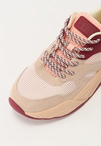 Light pink and beige sneaker with mesh and suede upper, maroon accents, patterned laces, and a textured rubber sole. Prominent brand tag visible.