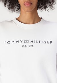 Bijela dukserica s natpisom "TOMMY HILFIGER" u crnoj boji, s "EST. 1985" ispod. Mekani materijal s okruglim izrezom i rebrastim rubovima.
