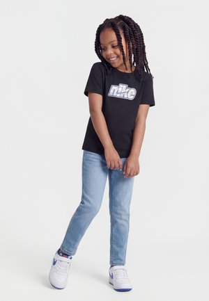 Jeune fille aux cheveux tressés portant un t-shirt Nike noir, un jean bleu clair et des baskets blanches Nike, souriante et regardant vers le bas.