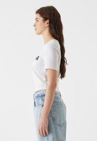 Camiseta blanca de manga corta con texto gráfico negro, combinada con unos vaqueros de denim de cintura alta en azul claro. Diseño sencillo, corte relajado, textura mínima.
