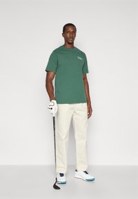 Πράσινο βαμβακερό μπλουζάκι κοντομάνικο, κρεμ παντελόνι, λευκά παπούτσια golf με μπλε λεπτομέρειες, κρατώντας μια ράβδο golf, με ένα λευκό γάντι.