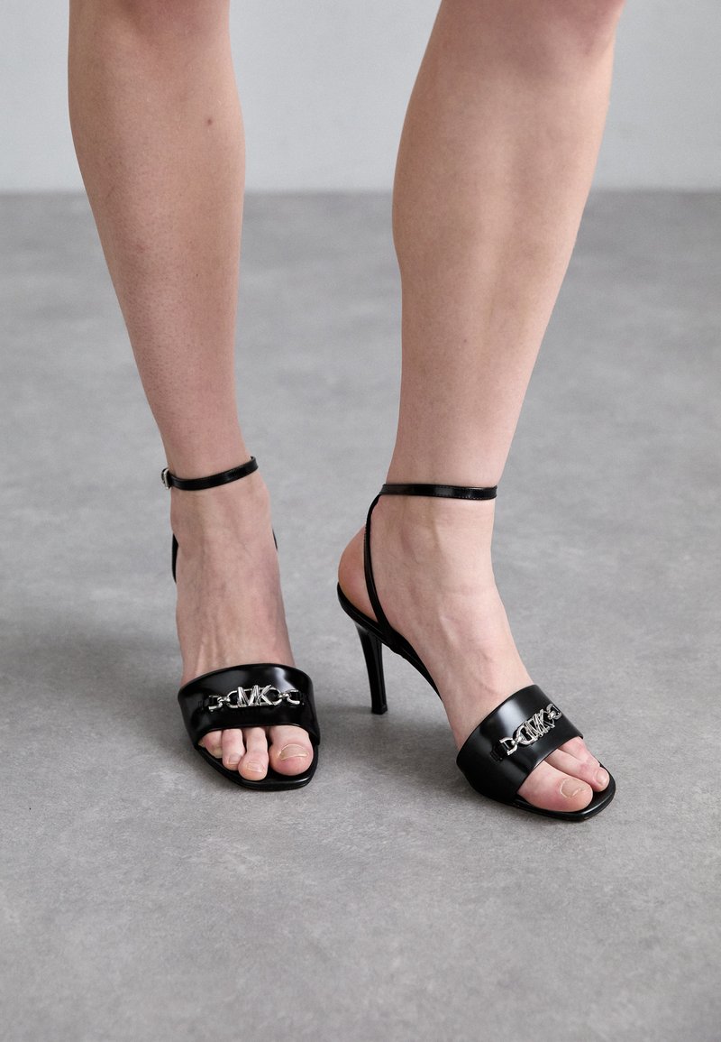 Black high-heeled sandals feature a smooth leather upper, toe straps with a silver chain detail, and a slender ankle strap.