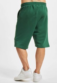 Shorts en coton verts avec une ceinture élastique, arrivant au genou, dotés de poches latérales et d'un détail logo sur la jambe gauche. Portés avec des baskets blanches.