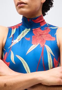 Άτομο που φοράει μπλε αμάνικη μπλούζα με κόκκινο φλοράλ μοτίβο και φερμουάρ μπροστά, με τα χέρια σταυρωμένα στο στήθος.