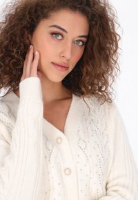 Kermakujainen neuletakki, jossa on köysineulekuvio, suuri nappikiinnitys ja koristeltu pienillä, värikkäillä helmillä ympäriinsä.