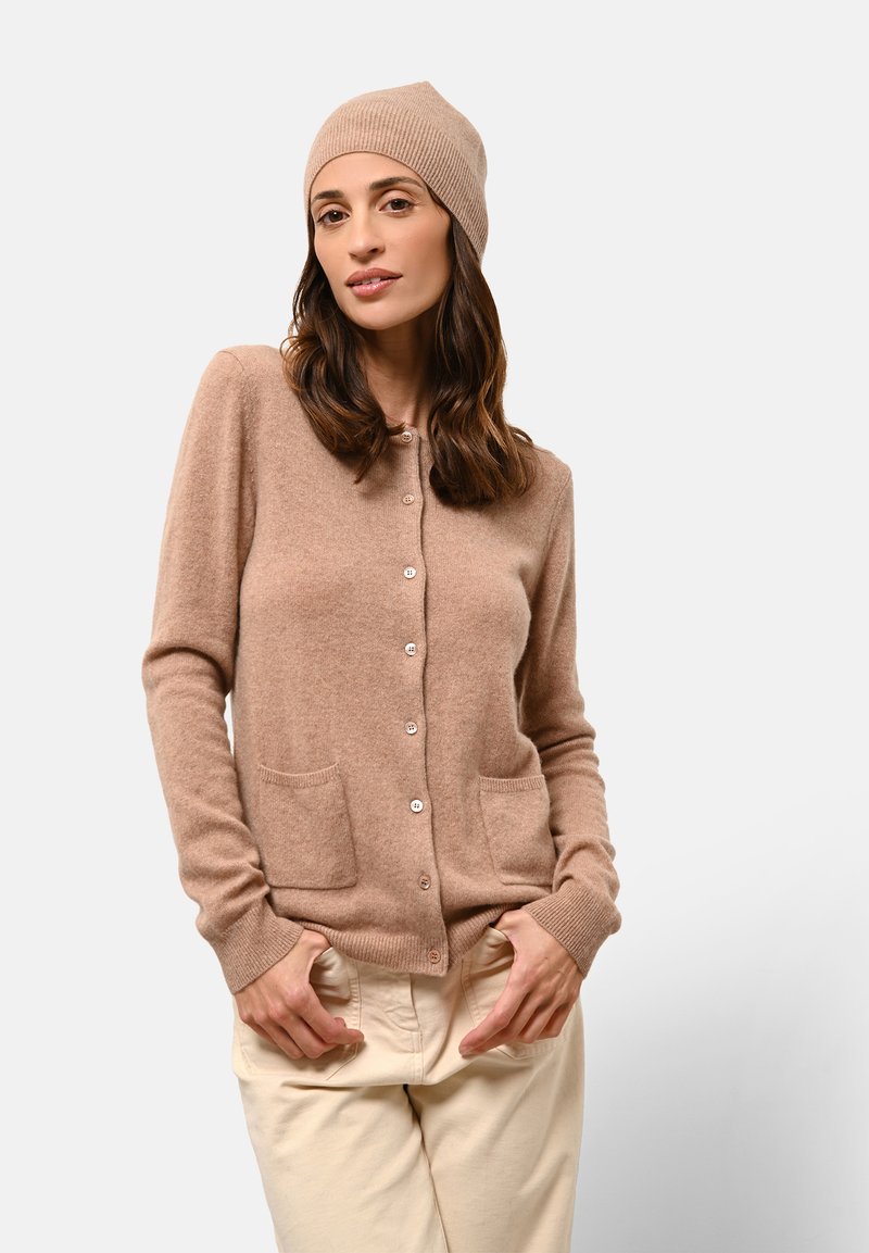 Cardigan in lana beige con scollo rotondo, chiusura a bottoni e due tasche frontali. Abbinato a un berretto a coste coordinato.