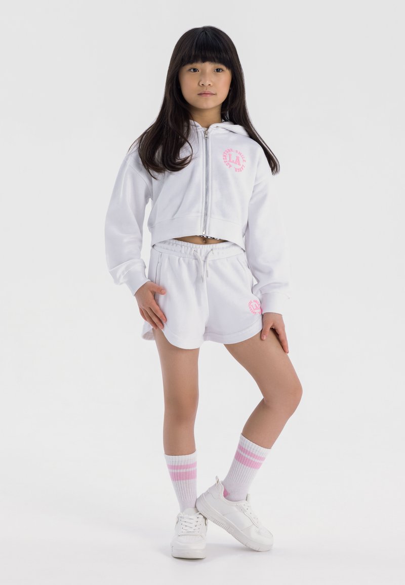 Felpa bianca corta con logo rosa, pantaloni corti bianchi abbinati, scarpe da ginnastica bianche e calze bianche con righe rosa, caratterizzate da una vestibilità rilassata e coulisse.