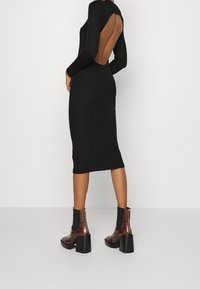 Schwarzes geripptes Strickkleid mit einem Rückenausschnitt, figurbetontem Schnitt und langen Ärmeln. Kombiniert mit braunen stöckeligen Ankle Boots mit derben Sohlen.