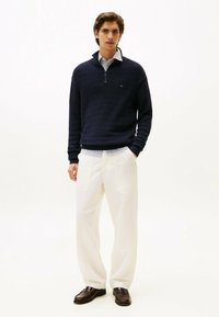 Maglione zip-up Navy con righe testurizzate, camicia bianca con colletto, pantaloni beige a gamba larga e mocassini marroni scuri. Design minimalista.