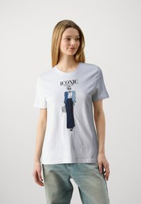 Witte katoenen T-shirt met een gedrukte illustratie van een figuur in een blauwe blazer en een lange donkere rok, met de tekst "ICONIC" er boven.