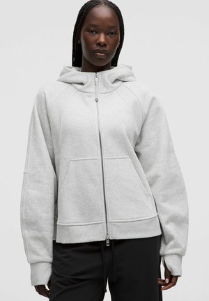 Frau trägt einen hellgrauen Hoodie mit Reißverschluss, Kapuze und Kängurutaschen, kombiniert mit schwarzen Hosen, stehend vor einem einfarbigen Hintergrund.