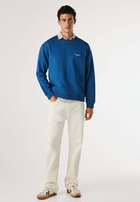 Giovane uomo con un felpa blu sopra una maglietta a righe, pantaloni color crema e sneakers bianche, in piedi contro uno sfondo chiaro e uniforme.