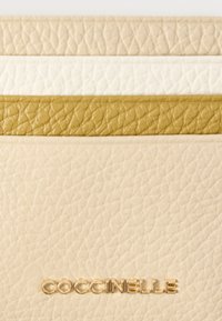 Δερμάτινη θήκη καρτών σε μπεζ, κρεμ και ελιά με υφή φινιρίσματος. Διαθέτει ανάγλυφο λογότυπο της μάρκας σε χρυσό στο κάτω μέρος.