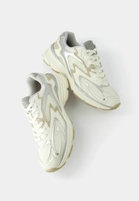 Sneakers featuring a light-colored mesh upper with silver and beige accents, thick sole, and zigzag design elements. Laces and padded collar.