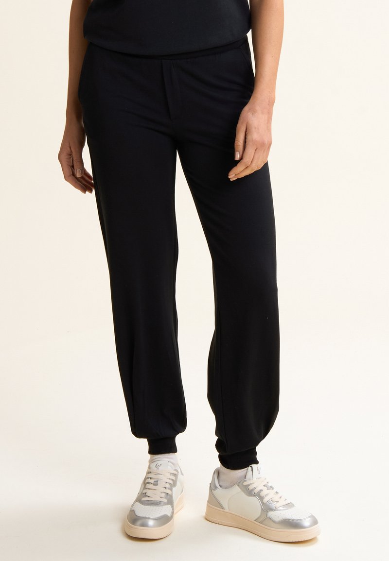 Jogging noir fuselé avec taille élastique et poignets; fabriqué en tissu doux. Associé à des baskets grises et argentées pour un look décontracté.