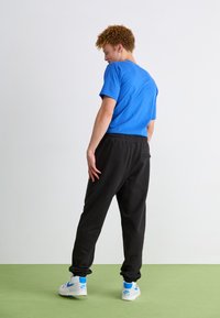 Camiseta de manga corta azul, pantalones deportivos negros con cintura elástica y zapatillas blancas con detalles azules, vistas desde atrás.