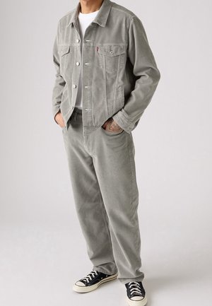 Mann trägt hellgraue Cordjacke und Hose mit weißem T-Shirt und schwarzen Sneakern, steht mit den Händen in den Taschen.