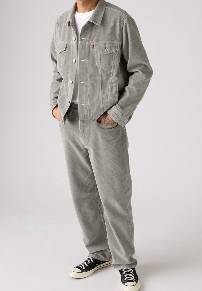 Homme portant une veste et un pantalon en velours côtelé gris clair, un t-shirt blanc et des baskets noires, debout les mains dans les poches.