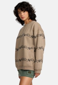 Felpa beige con stampe floreali nere e una mano sul petto. Presenta un scollo a girocollo e spalle cadenti. Abbinata a pantaloni corti verdi.