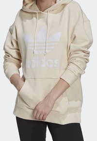 Krémově zbarvená mikina s kapucí z měkké látky, zdobená velkým bílým logem Adidas, stahovací kapucí a přední kapsou ve stylu klokánka.