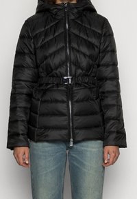 Giubbotto puffer nero con design trapuntato, cintura in vita cincherata e chiusura con zip. Presenta un cappuccio e maniche lunghe.