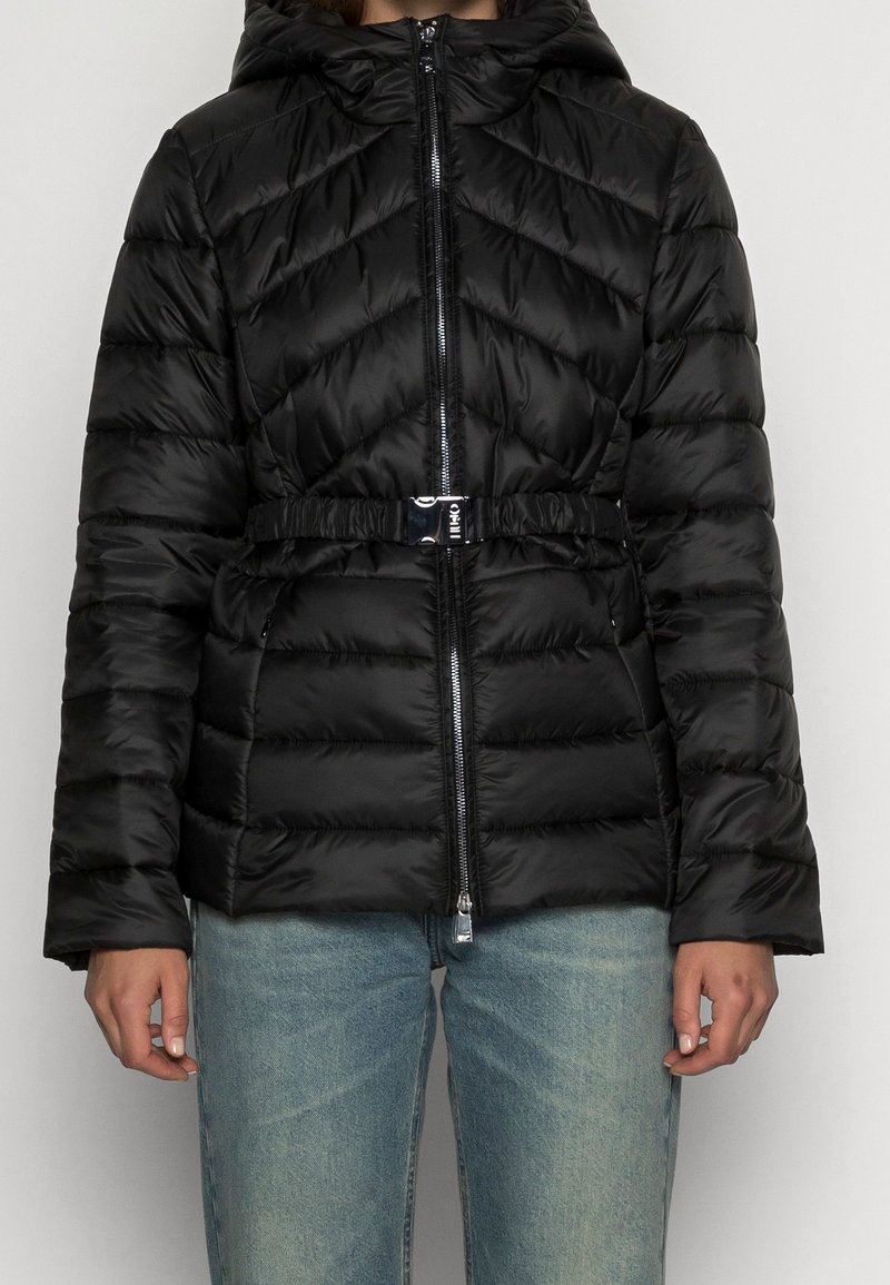 Giubbotto puffer nero con design trapuntato, cintura in vita cincherata e chiusura con zip. Presenta un cappuccio e maniche lunghe.