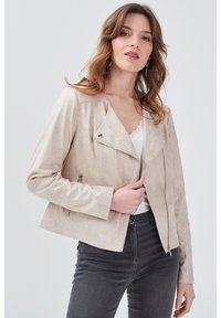 Veste en suède synthétique beige avec une fermeture éclair asymétrique, des manches longues, des poches latérales et une silhouette ajustée. Portée par-dessus un haut blanc à dentelle.