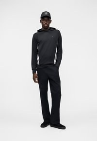 Camisola preta com riscas laterais brancas, combinada com calças pretas e ténis. Um boné preto apresenta um logótipo. O conjunto é simples e moderno.