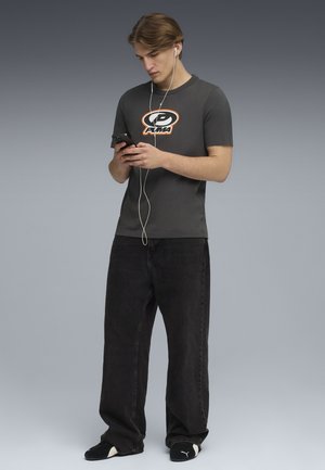 Grå T-shirt med orange Puma-logo, parret med sorte breddejeans og sorte slippers. Personen holder en smartphone og har høretelefoner på.