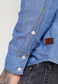 Lichtblauwe denim shirtmouw met witte knopen en contrasterend stiksel. Bevat een leren logo-patch in bruin nabij de manchet.