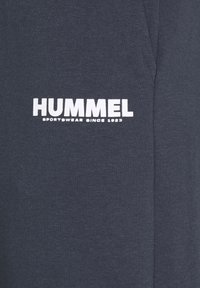 Tamsiai mėlynos sportinės šortai su lygia tekstūra, su ryškiu baltu "HUMMEL" logotipu ir tekstu "SPORTSWEAR SINCE 1923."