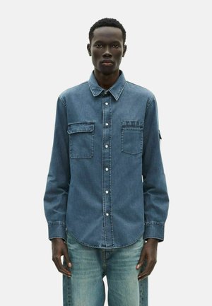Blaue Jeanshemd mit durchgehender Knopfleiste, zwei Brusttaschen und spitzem Kragen. Lange Ärmel mit Knopfmanschetten. Abgenutzte Textur.