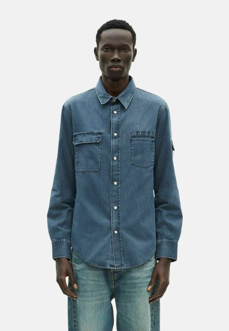 Camicia in denim blu con chiusura a bottoni, due tasche sul petto e colletto a punta. Maniche lunghe con polsini abbottonati. Texture vissuta.