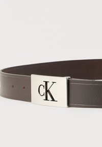 Σκούρο καφέ δερμάτινη ζώνη με ορθογώνια ασημί αγκράφα που διαθέτει το κομμένο λογότυπο "cK".
