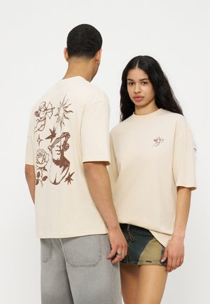 Dos modelos llevan camisetas beige de gran tamaño; una tiene un diseño de querubín y sol en la espalda, la otra presenta un pequeño diseño de querubín en el frente.