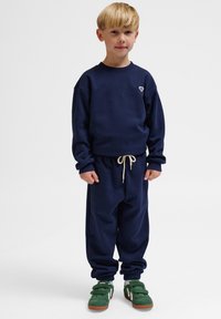 Granatowy zestaw bluzy i spodni dresowych z elastycznymi mankietami i sznurkiem w talii. Zawiera małe logo na piersi; połączony z zielonymi trampkami.
