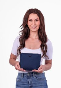 Námořnický látkový sáček s měkkou texturou, se zipovým uzávěrem a černým logem na přední straně; drží ho osoba v neformálním oblečení.