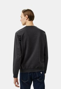 Felpa nera con maniche lunghe, scollatura rotonda, polsini e orlo a costine, realizzata in tessuto morbido. Indossata con jeans blu caratterizzati da un logo triangolare.