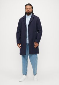 Cappotto in lana blu navy con chiusura a bottoni, collo con revers e tasche laterali, indossato sopra una polo azzurro chiaro e jeans larghi. Sneakers bianche.