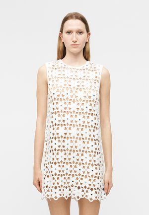 Žena oblečená v bielych bezrukávových šatách z háčkovanej tkaniny s kvetinovými vzormi, stojaca pred jednoduchým svetlým pozadím, otočená čelom.